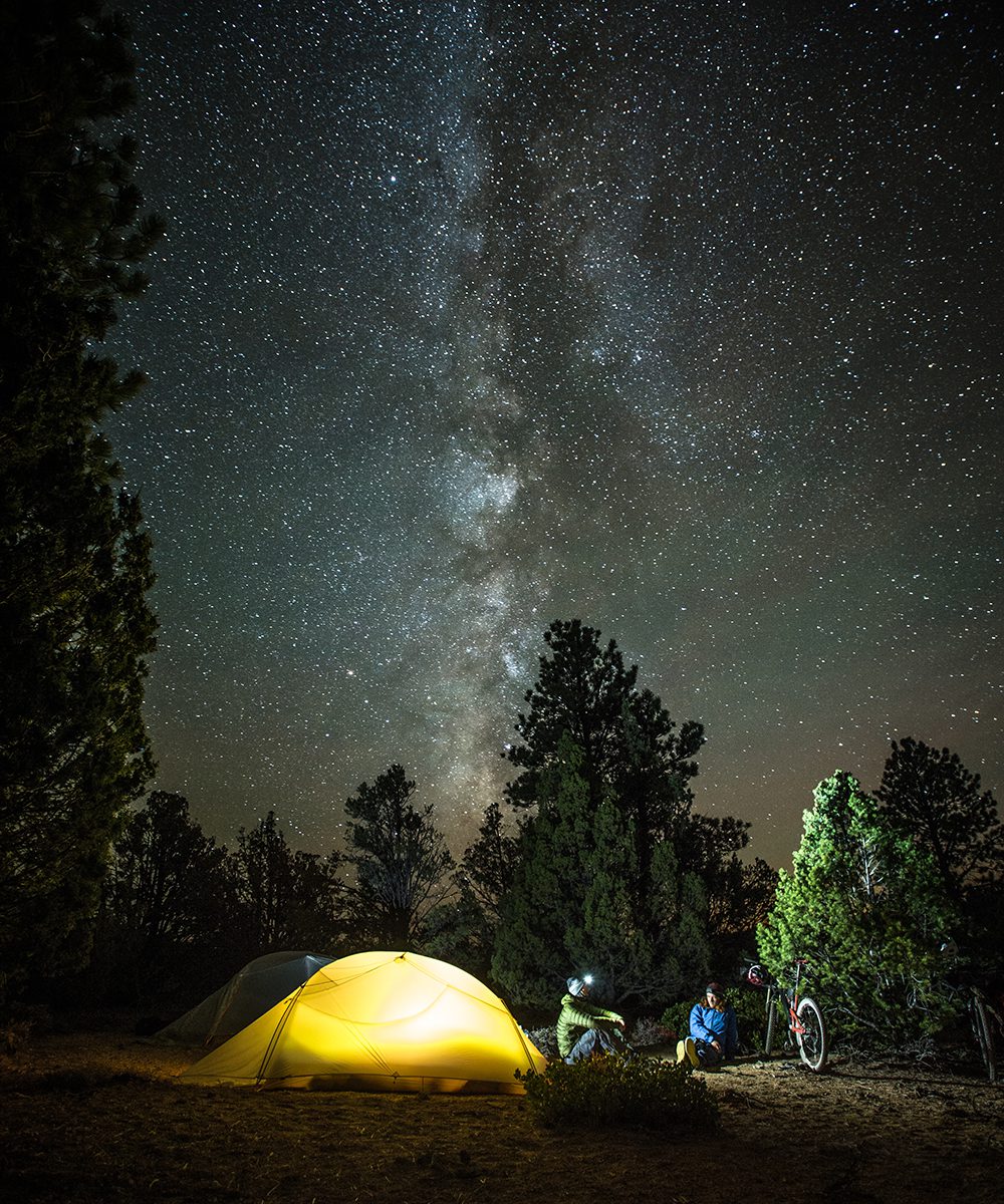 Eric Porter and Kurt Gensheimer sitting outside their glowing tent as the Milky Way up in the night sky near Pole Canyon in Southern Utah, by Scott Markewitz